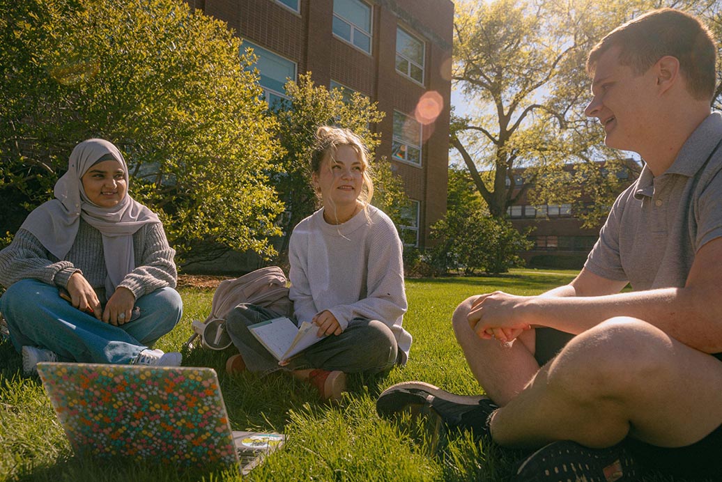 Three students sit on the grass around a laptop.