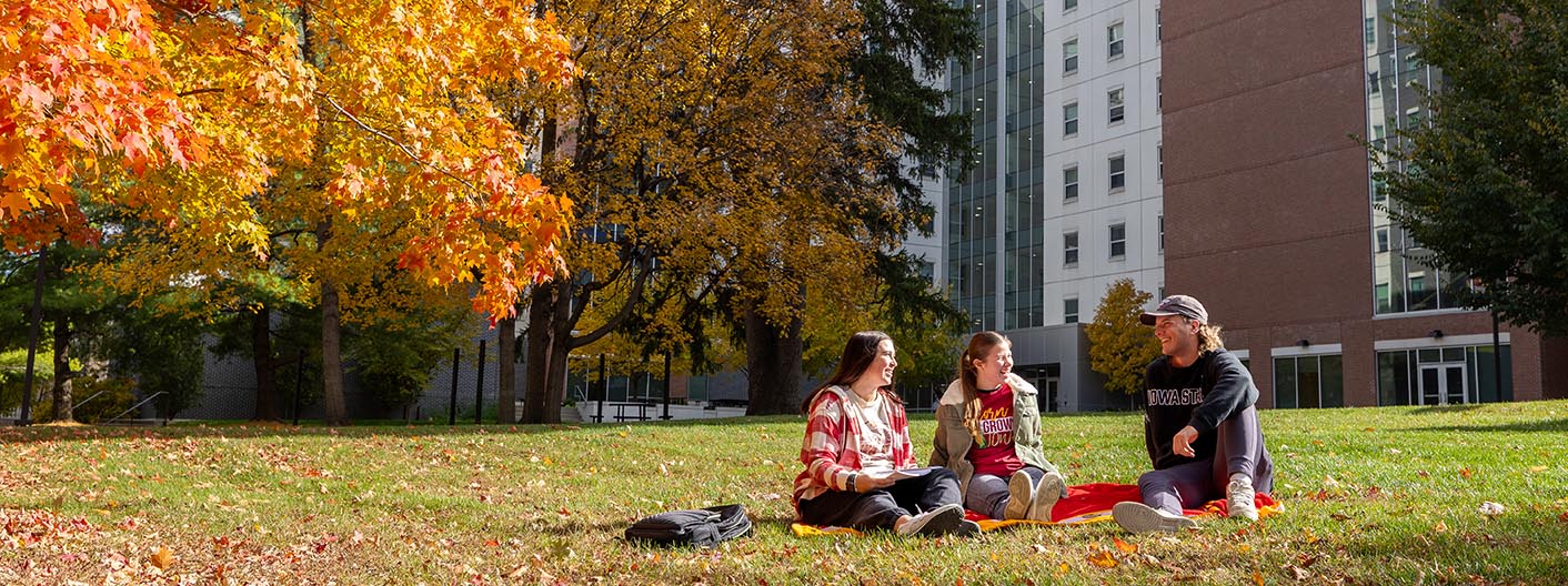 Three students sit on the grass outside of Geoffroy Hall on a fall day.